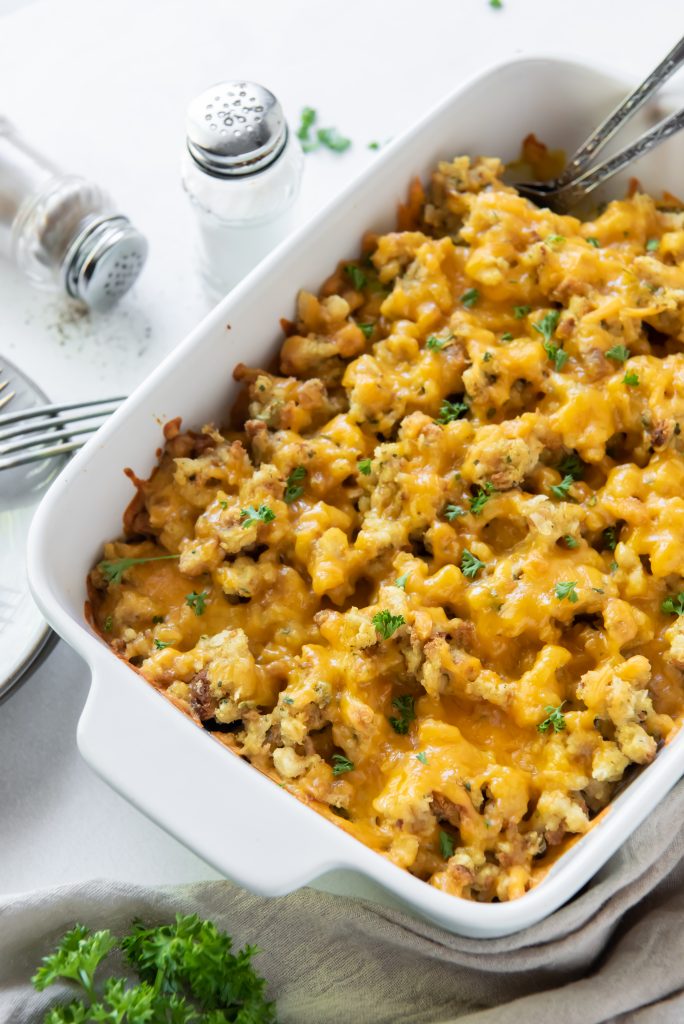 A baking dish brimming with cheesy chicken broccoli stuffing casserole is topped with chopped herbs. Beside it, salt and pepper shakers, a fork, and a folded cloth await. Fresh parsley is scattered on the table for garnish.