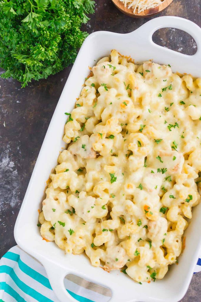 A white casserole dish brimming with a delightful mac and cheese, topped with melted cheese and sprinkled with parsley, reminiscent of a classic chicken alfredo pasta bake. Resting on a dark surface beside fresh parsley and a striped cloth, its an inviting culinary masterpiece.