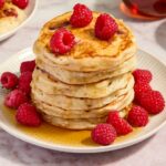 a stack of pancakes on a white plate with raspberries and syrup