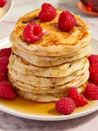 a stack of pancakes on a white plate with raspberries and syrup