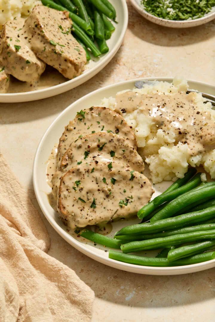 pork tenderloin on a white plate with cream sauce, mashed potatoes, and green beans