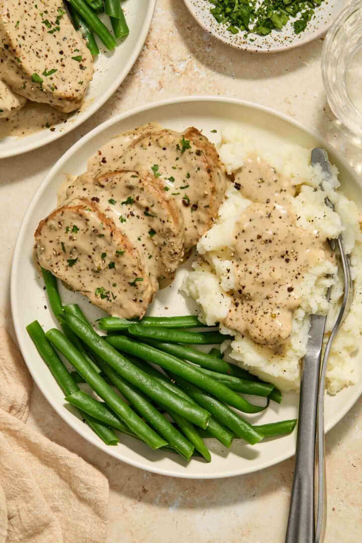 pork tenderloin with cream sauce on a white plate with mashed potatoes and green beans