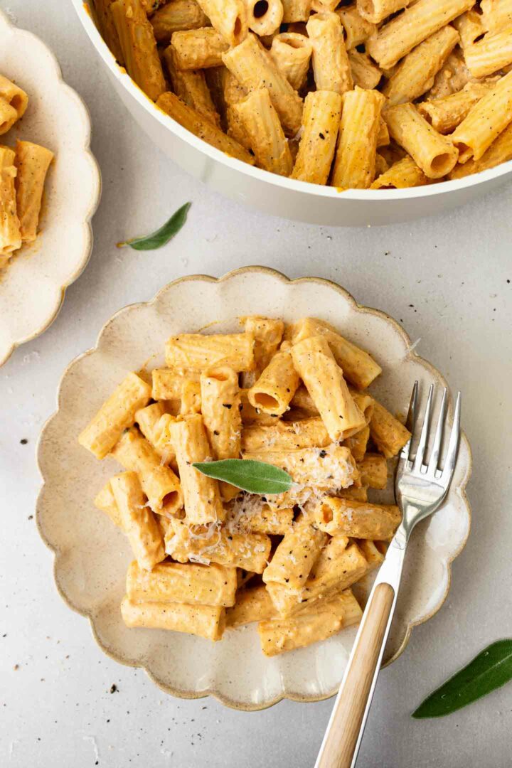 pumpkin alfredo pasta on white plate with a silver fork next to another plate of pasta and rest of pasta in white skillet