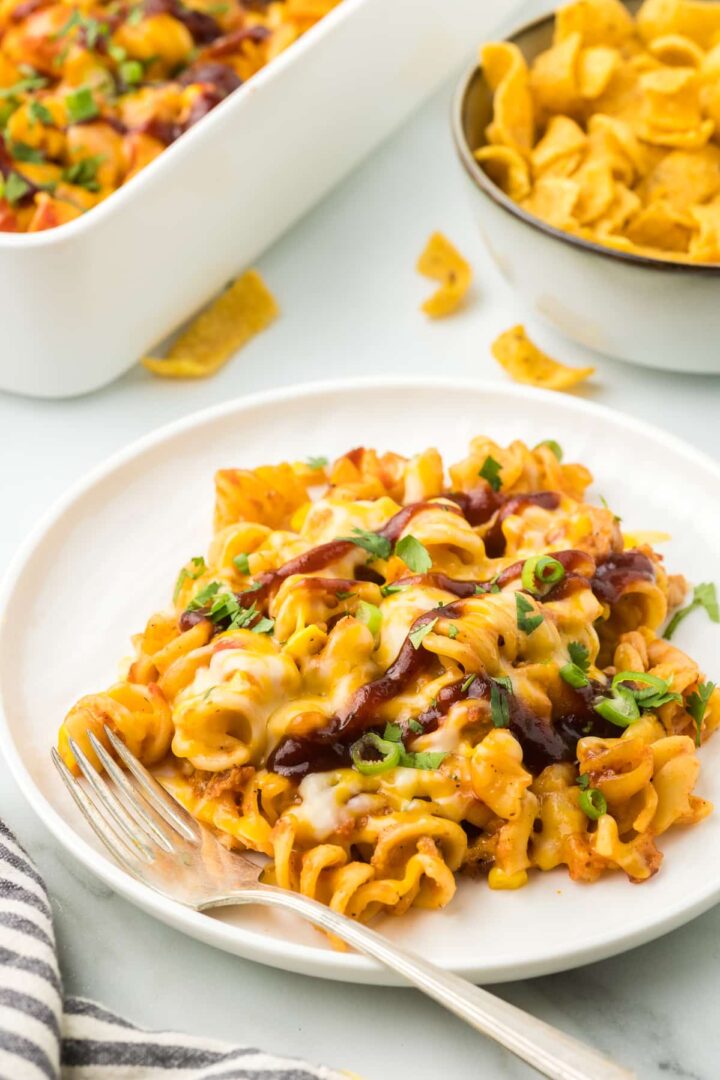 A plate of cheesy pasta topped with barbecue sauce and chopped green onions is in the foreground. A fork rests on the plate. In the background, there is a bowl of corn chips and a baking dish with more pasta. A striped napkin is nearby.