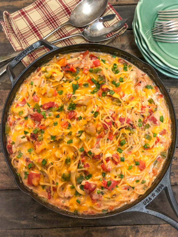 A skillet filled with baked cheesy spaghetti, featuring pasta, diced tomatoes, green onions, and melted cheese. A folded checkered napkin with utensils is beside the skillet on a rustic wooden table.