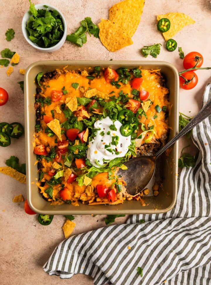 A square pan filled with a taco casserole topped with melted cheese, chopped tomatoes, jalapeños, cilantro, and a dollop of sour cream. A spoon rests inside the pan. Tortilla chips and cherry tomatoes are scattered around.