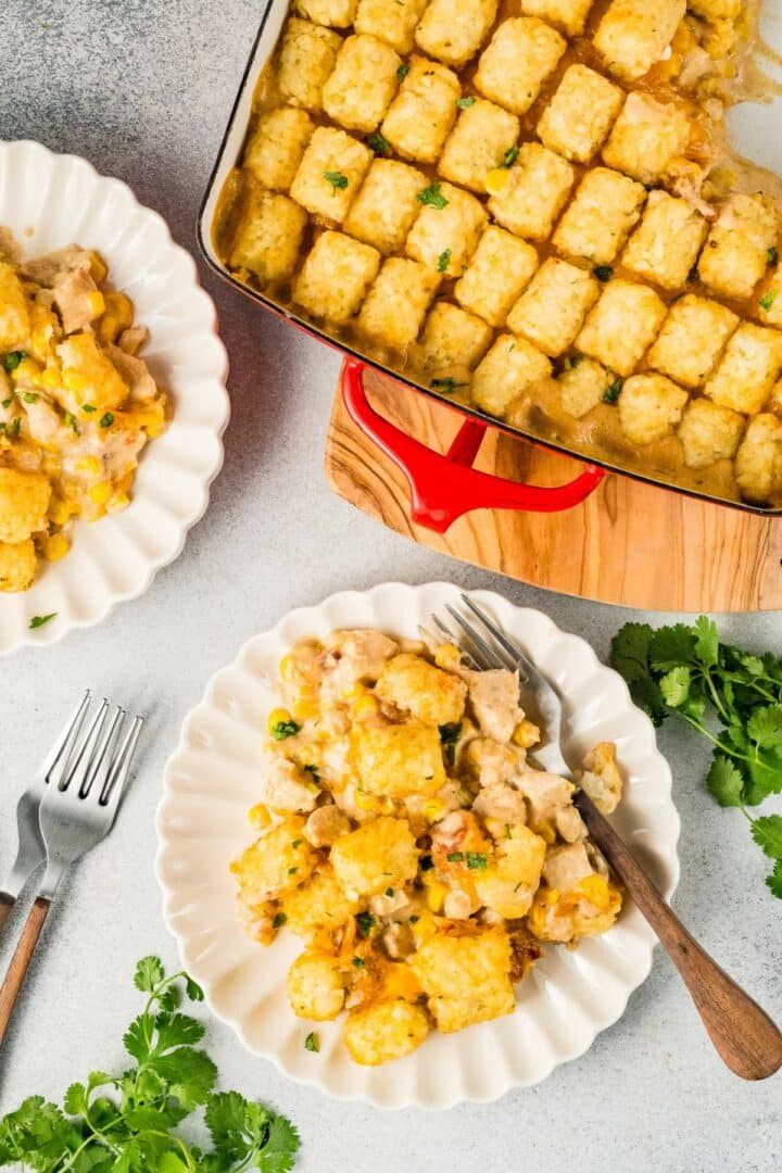 A casserole dish filled with crispy tater tots on top of a creamy casserole sits on a wooden board. Two plates with servings of the casserole are nearby, garnished with herbs. Forks are placed beside the plates. Fresh cilantro decorates the scene.