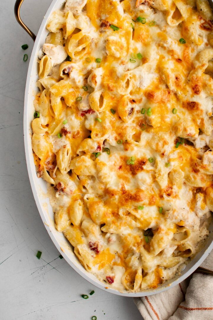 A baked pasta dish with melted cheese and browned top filling an oval white casserole dish. Small pieces of green garnish are sprinkled over the cheese. A striped cloth is partially visible underneath the dish.