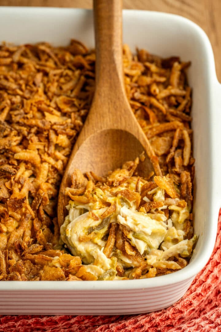 A white casserole dish filled with crispy baked topping and a creamy mixture beneath. A wooden spoon rests in the dish, scooping up some of the creamy filling. The dish is placed on a red textured cloth.