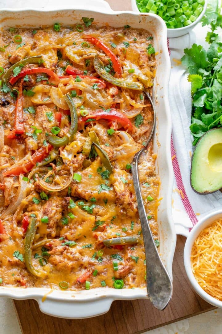 A casserole dish filled with cheesy chicken and vegetable bake, including red and green bell peppers and onions. A spoon is in the dish. Next to it are chopped green onions, cilantro, sliced avocado, and shredded cheese in bowls.