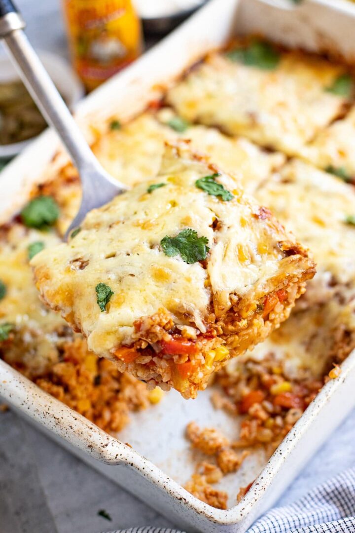 A square slice of cheesy baked casserole with quinoa, vegetables, and herbs is lifted from a baking dish. The dish is garnished with cilantro and appears hot and freshly cooked.