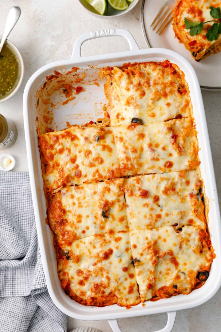 A white rectangular baking dish filled with cheesy enchiladas, topped with melted cheese and sliced into squares. A portion is missing from one corner. Next to the dish are lime wedges, a fork, and a small bowl of green sauce.