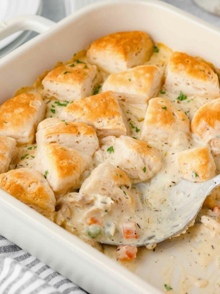 A baking dish filled with a creamy chicken casserole, topped with golden-brown biscuit pieces. The dish is garnished with herbs and includes visible pieces of vegetables like carrots and peas. A serving utensil is partially submerged in the casserole.