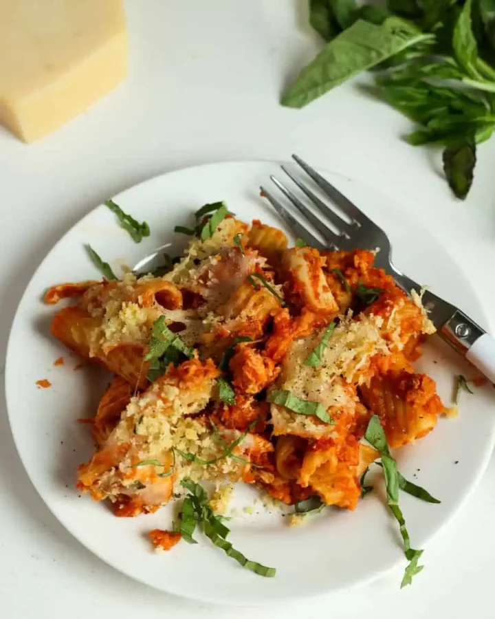 A white plate with baked ziti covered in tomato sauce, melted cheese, and sprinkled with chopped basil. A fork rests on the plate. Fresh basil leaves and a block of cheese are nearby on the white surface.