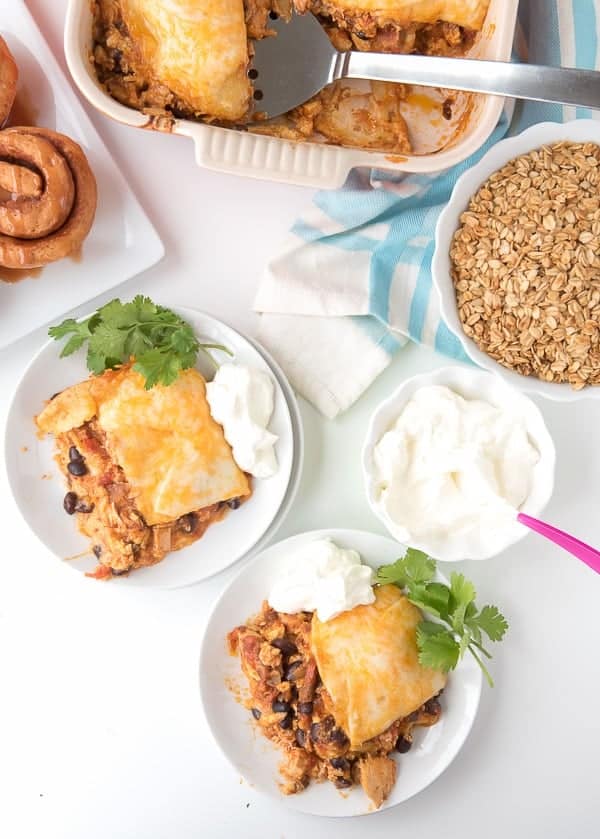 Two plates with a cheesy baked casserole topped with cilantro and sour cream. Next to them, a baking dish with more casserole, a bowl of sour cream, a plate of cinnamon rolls, and a bowl of granola, all on a white surface with a blue-striped cloth.