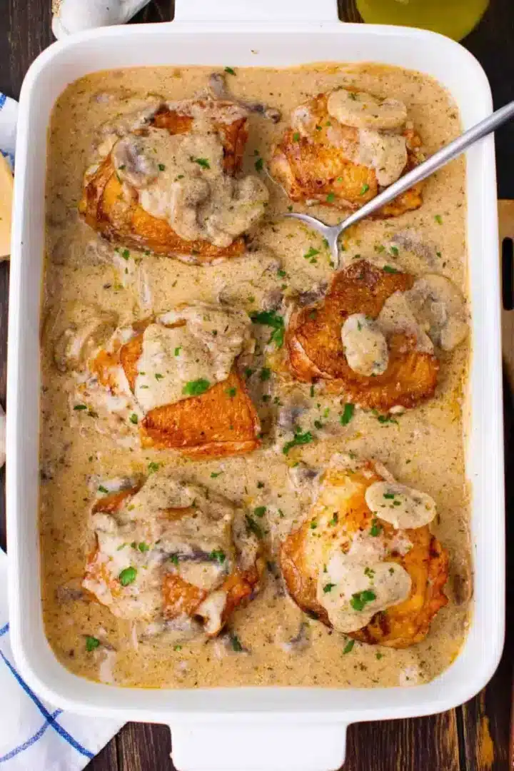 A white baking dish filled with roasted chicken pieces in a creamy mushroom sauce, garnished with chopped parsley. A serving spoon rests in the dish. The sauce is rich with visible slices of mushrooms.