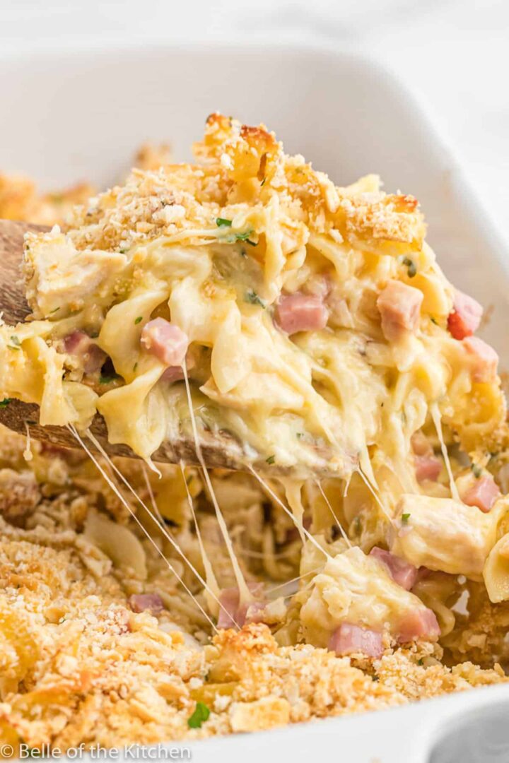 A close-up of a creamy pasta bake being scooped from a dish. The pasta is covered in a melted cheese sauce with pieces of ham and sprinkled with a crispy breadcrumb topping.